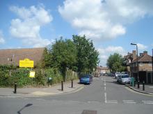 A street scene with houses on both sides, a few parked cars, and a yellow sign on the left. The sky is partly cloudy