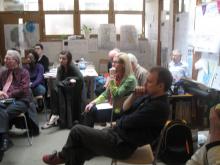 A group of people seated in a classroom-style setting, attentively listening. Posters and papers are visible on the walls in the background