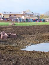 A muddy field with a small pond and scattered rocks, with a building and graffiti-covered wall in the background