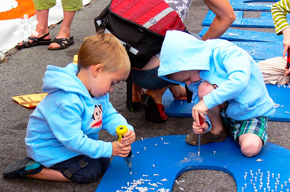 Two young children in blue hoodies crouch on the ground, engaged in a hands-on activity with screwdrivers on blue boards. They are surrounded by adults and kids, participating in a creative outdoor event on a sunny day