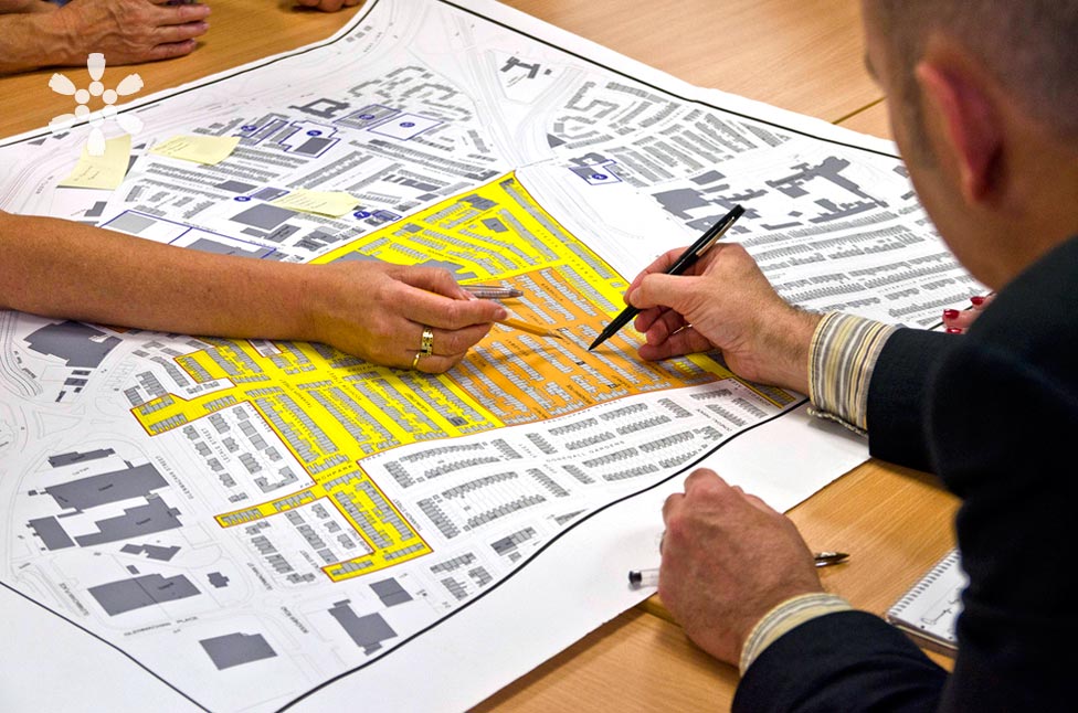 People reviewing a detailed, color-coded city planning map on a table. One persons hand is pointing with a pen, while another uses a marker to highlight areas. The map shows streets, buildings, and various marked zones