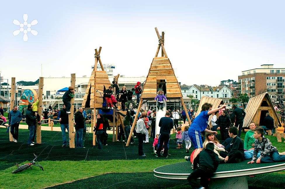 A busy playground with various wooden climbing structures and slides. Children are playing, climbing, and socializing while adults supervise. Buildings and a clear sky are in the background